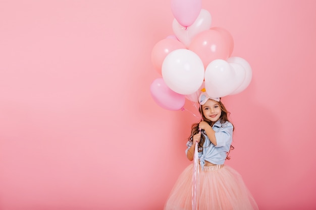 Feliz celebración de la fiesta de cumpleaños con globos voladores de encantadora niña linda en falda de tul sonriendo a cámara aislada sobre fondo rosa. Sonrisa encantadora, que expresa felicidad. Lugar para el texto