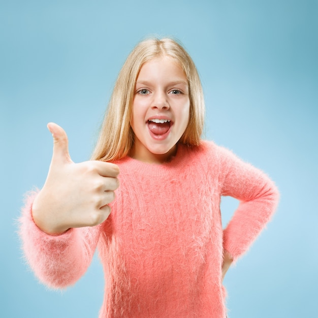 Feliz chica adolescente de pie, sonriendo aislado sobre fondo de estudio azul de moda. Hermoso retrato femenino. Los jóvenes satisfacen a la chica con el signo ok. Las emociones humanas, el concepto de expresión facial. Vista frontal.