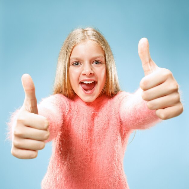 Feliz chica adolescente de pie, sonriendo aislado sobre fondo de estudio azul de moda. Hermoso retrato femenino. Los jóvenes satisfacen a la chica con el signo ok.