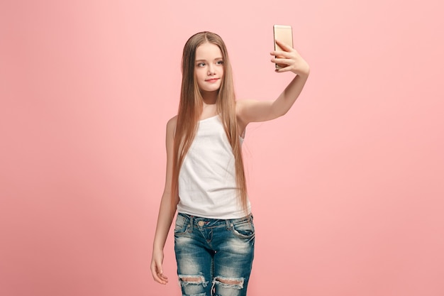 Feliz chica adolescente de pie, sonriendo en la pared rosa, haciendo foto selfie por teléfono móvil. Las emociones humanas, el concepto de expresión facial. Vista frontal.