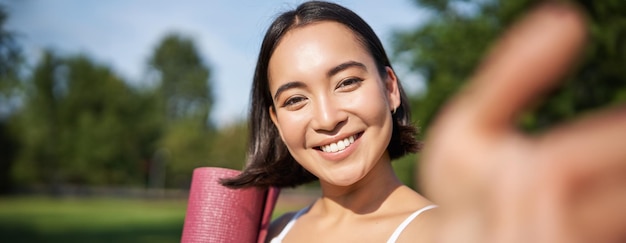 Feliz chica asiática de fitness se toma selfie con una estera de yoga de goma en el parque, una joven deportista sana hace