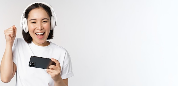 Feliz chica asiática jugadora jugando en el teléfono móvil mirando en el teléfono inteligente con auriculares de pie sobre fondo blanco.