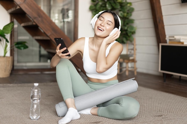 Feliz chica asiática en ropa de fitness sentado en casa en el suelo escuchando música y ejercicio sosteniendo sm ...