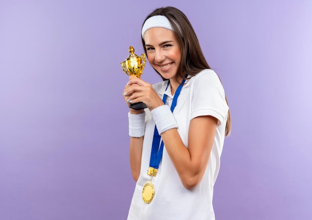 Feliz chica bastante deportiva con diadema y muñequera y medalla sosteniendo la taza aislada en la pared púrpura con espacio de copia