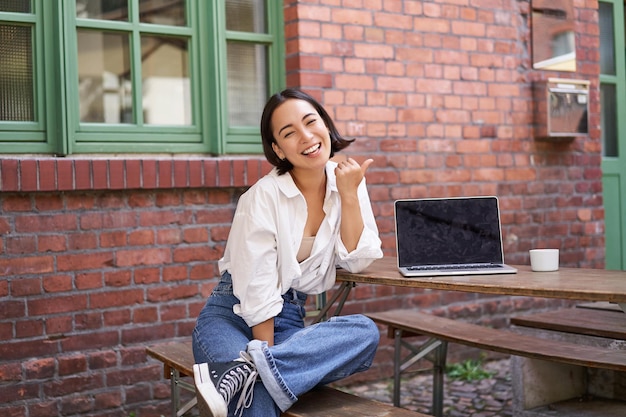 Foto gratuita feliz chica coreana con computadora portátil sentada en un café riéndose y señalando el espacio de copia en la pared de ladrillo