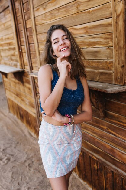 Feliz chica delgada en falda de moda y camiseta azul riendo y tocando su barbilla.