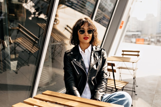 Feliz chica elegante con pelo corto con gafas y chaqueta de cuero con labios rojos descansando en la terraza de verano en la luz del sol