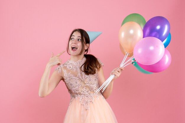 Feliz chica fiestera con gorra de fiesta sosteniendo globos apuntando con el dedo su sonrisa en rosa