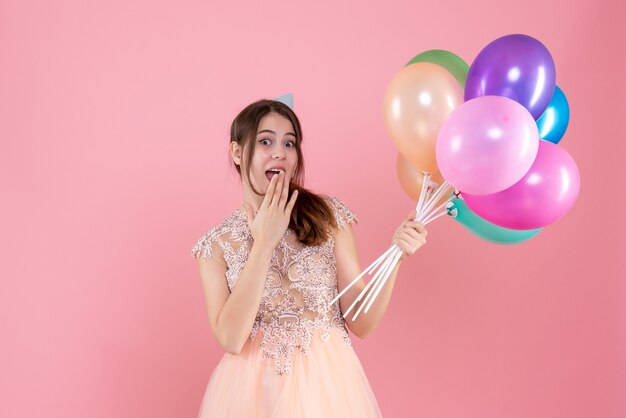 Feliz chica fiestera con gorro de fiesta sosteniendo globos poniendo la mano a la boca en rosa