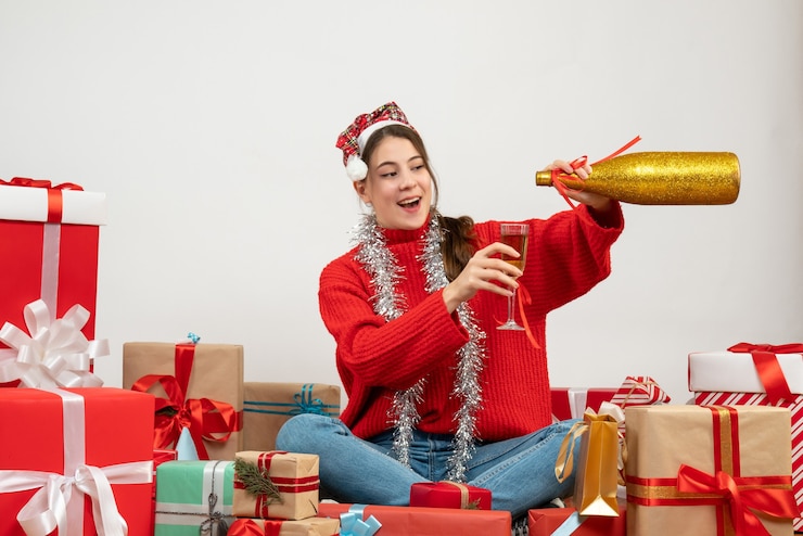 Foto gratuito feliz chica fiestera con gorro de papá noel vertiendo champán sentados alrededor presenta en blanco