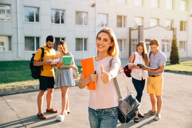 Feliz chica hermosa con cuadernos y mochila mostrando pulgar arriba y sonriendo