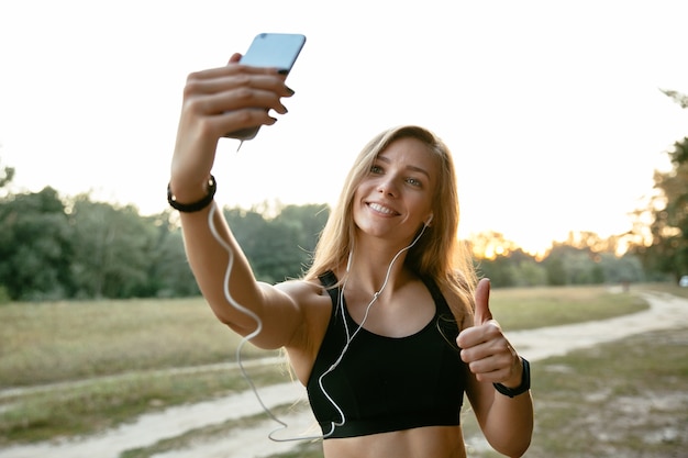 Feliz chica increíble en auriculares, toma un selfie en teléfono móvil, mostrando un pulgar arriba