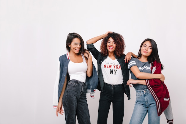 Feliz chica latina en jeans divirtiéndose con compañeros de la universidad posando con una sonrisa. Retrato de interior de una hermosa dama africana de pie entre amigos y jugando con el pelo rizado.