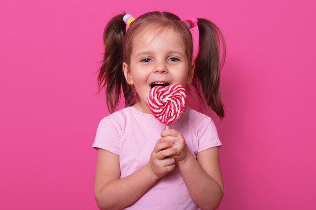 Feliz chica linda viste camiseta rosa, se encuentra aislado en rosa, tiene paleta brillante en las manos. Niño alegre con la boca abierta cata deliciosos dulces. Concepto de infancia y gustos.