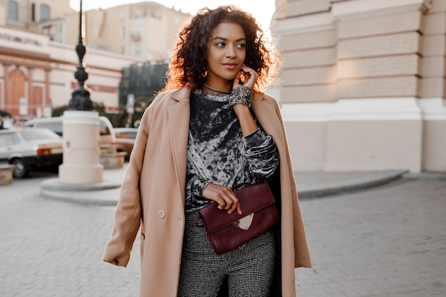 Feliz chica negra con sonrisa sincera en suéter de terciopelo gris increíble, abrigo de lana beige, accesorios de joyería de lujo caminando en parís cerca del teatro.