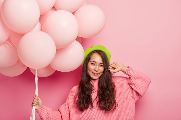 Feliz chica oriental de pelo oscuro sostiene un montón de globos de aire, viste boina verde y suéter de gran tamaño