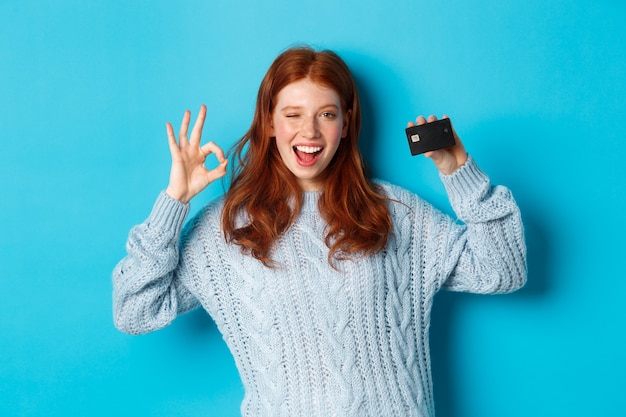 Feliz chica pelirroja en suéter mostrando tarjeta de crédito y signo bien, recomendando oferta bancaria, de pie sobre fondo azul.