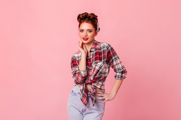 Feliz chica pinup sonriendo en el espacio rosa. Disparo de estudio de elegante mujer jengibre en jeans y camisa a cuadros.