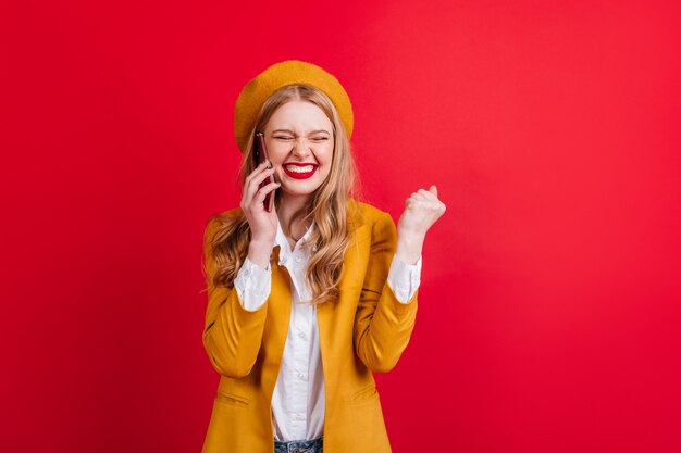 Feliz chica rubia en boina hablando por teléfono con una sonrisa. mujer francesa emocionada aislada en la pared roja.