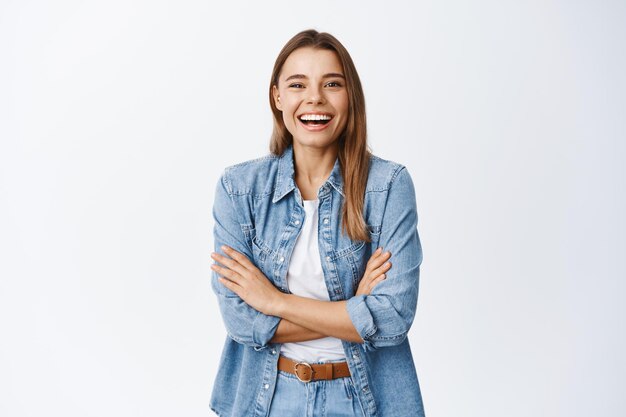 Feliz chica rubia en ropa casual, riendo de una broma divertida, luciendo despreocupada y divirtiéndose, de pie con los brazos cruzados sobre el pecho, blanco