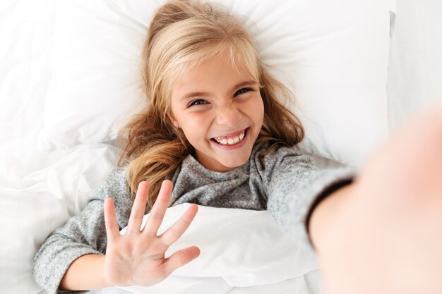 Feliz chica rubia sonriente tomando selfie mientras está acostado en la cama