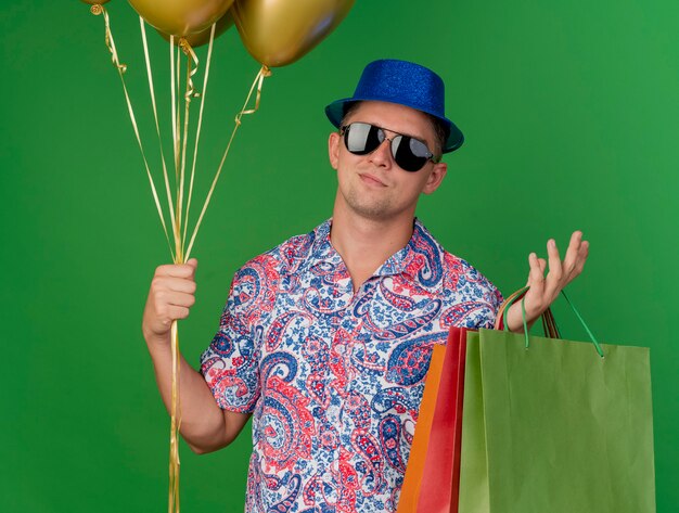 Feliz chico de fiesta joven con sombrero azul y gafas sosteniendo globos con bolsas de regalo y extendiendo la mano aislada sobre fondo verde