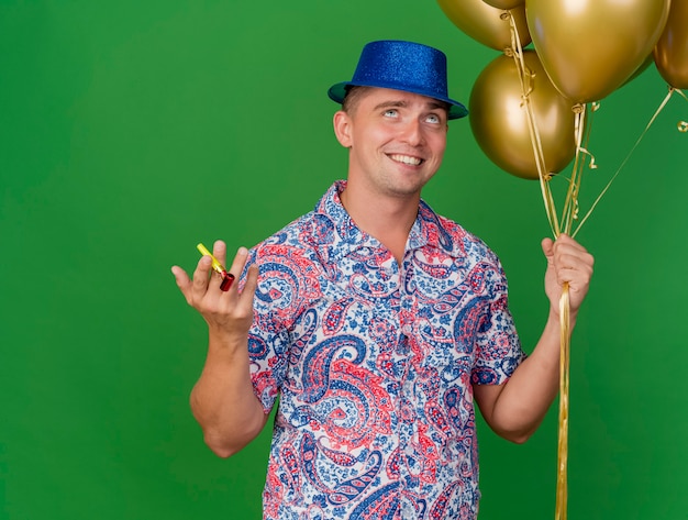 Feliz chico de fiesta joven con sombrero azul sosteniendo globos con soplador de fiesta aislado sobre fondo verde