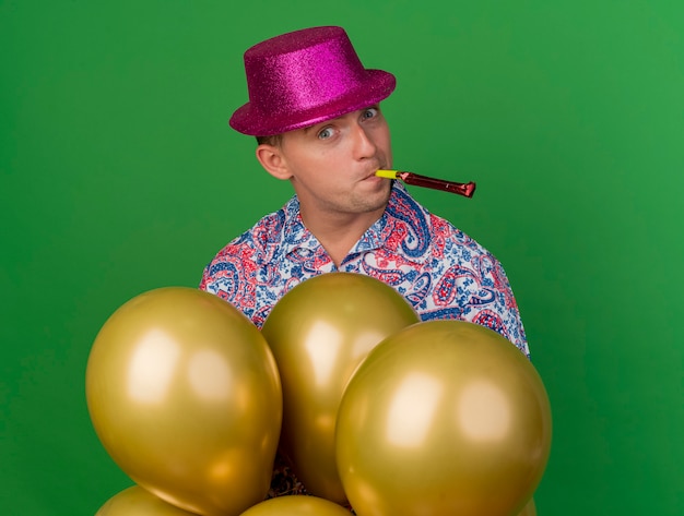 Feliz chico de fiesta joven con sombrero rosa de pie detrás de globos soplando soplador de fiesta aislado en verde