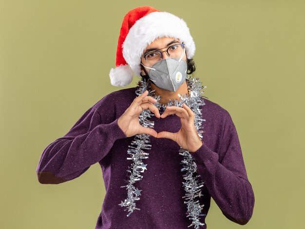 Feliz chico guapo joven con sombrero de Navidad y máscara médica con guirnalda en el cuello mostrando gesto de corazón aislado en la pared verde oliva