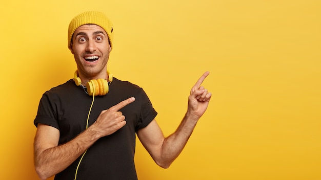 Foto gratuita feliz chico hipster apunta a un lado con ambos dedos, anuncia espacio de copia para su contenido publicitario