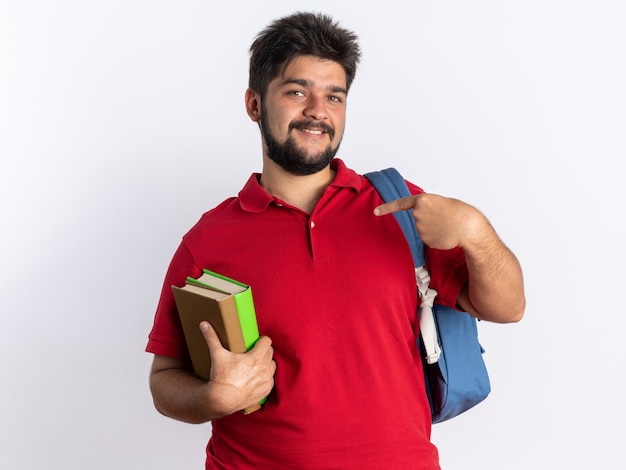 Feliz chico joven estudiante barbudo en polo rojo con mochila sosteniendo cuadernos apuntando con el dedo índice a cuadernos sonriendo alegremente de pie