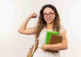 Foto gratuita feliz colegiala bonita joven con gafas y bolsa trasera sosteniendo libros gesticulando fuerte aislado en la pared blanca