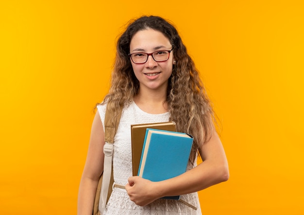 Feliz colegiala bonita joven con gafas y bolsa trasera sosteniendo libros mirando al frente aislado en la pared amarilla