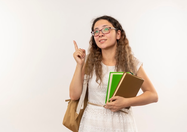 Feliz colegiala bonita joven con gafas y bolsa trasera sosteniendo libros mirando hacia arriba levantando el dedo aislado en la pared blanca