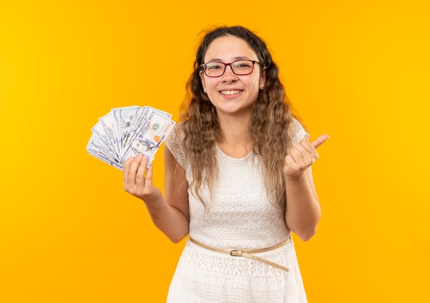 Feliz colegiala bonita joven con gafas sosteniendo dinero mostrando el pulgar hacia arriba aislado en amarillo con espacio de copia