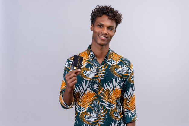 Feliz y complacido joven guapo de piel oscura con cabello rizado en hojas camisa estampada y mostrando tarjeta de crédito