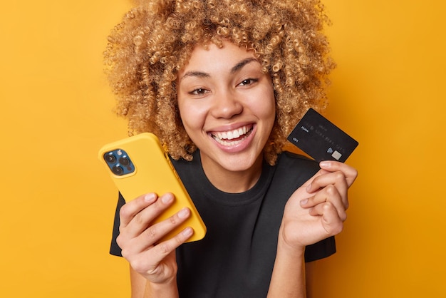 La feliz compradora rizada disfruta de pagar fácilmente los bienes en línea, las sonrisas muestran ampliamente los dientes blancos, usa el teléfono móvil y la tarjeta de crédito, vestida con una camiseta negra informal aislada sobre un fondo amarillo