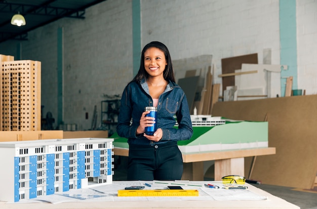 Feliz dama afroamericana con termo de pie cerca del modelo de edificio