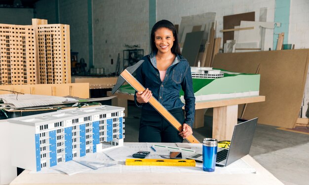 Feliz dama afroamericana con varita cerca de mesa con laptop y modelo de edificio