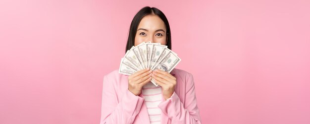 Feliz dama asiática en traje sosteniendo dólares de dinero con expresión de cara complacida de pie sobre fondo rosa