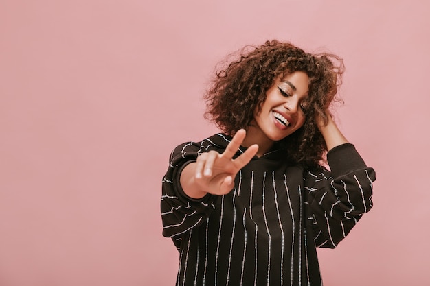 Feliz dama de pelo rizado con maquillaje fresco en ropa elegante a rayas mostrando el signo de la paz y sonriendo con los ojos cerrados en la pared rosa.