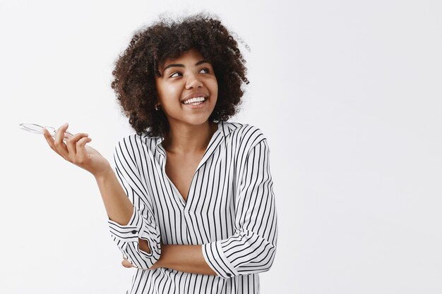 Feliz y despreocupada empresaria afroamericana moderna en blusa a rayas de moda sosteniendo vasos en la mano y mirando a la esquina superior derecha con una amplia sonrisa satisfecha