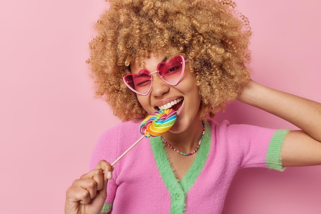 Foto gratuita una feliz y despreocupada mujer de cabello rizado no puede vivir sin dulces, dulces multicolores, lleva gafas de sol en forma de corazón y una camiseta aislada sobre un fondo rosa sabor arco iris concepto de vida dulce