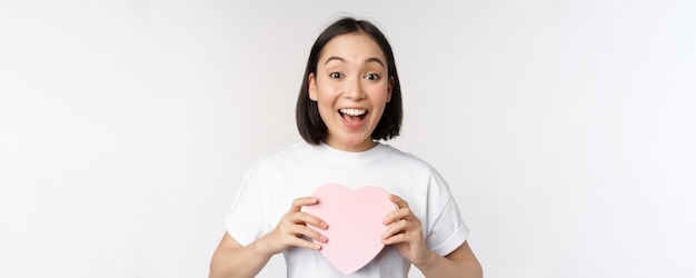 Feliz día de san valentín, una chica asiática recibe un regalo romántico con una caja en forma de corazón y una sonrisa emocionada.