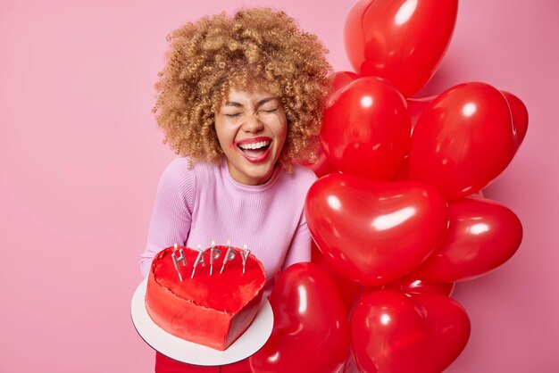 Feliz Día de San Valentín Una joven de cabello rizado y llena de alegría sostiene un delicioso pastel y un montón de globos inflados en forma de corazón expresa emociones positivas y prepara una sorpresa para su marido de vacaciones