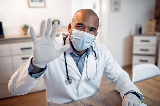 Feliz doctor negro con mascarilla saludando a alguien durante una conferencia telefónica desde su oficina