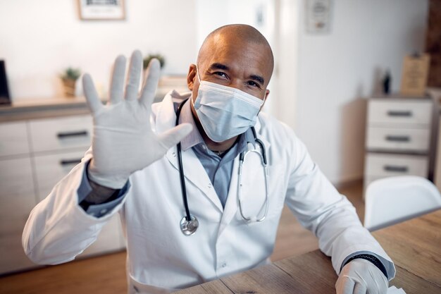 Feliz doctor negro con mascarilla saludando a alguien durante una conferencia telefónica desde su oficina