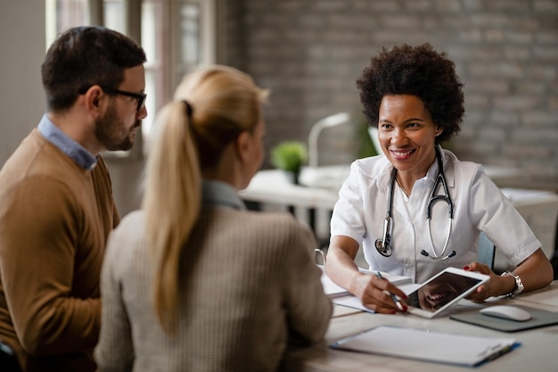 Feliz doctora negra hablando con una pareja y mostrándoles los resultados de las pruebas médicas en un panel táctil durante las consultas en la clínica