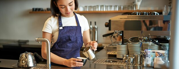 Feliz dueña de un café sonriente chica barista en delantal haciendo capuchino latte arte con leche al vapor
