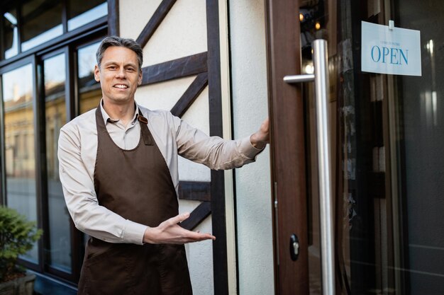 El feliz dueño del pub da la bienvenida a los invitados mientras está parado en la puerta de entrada y mira la cámara.